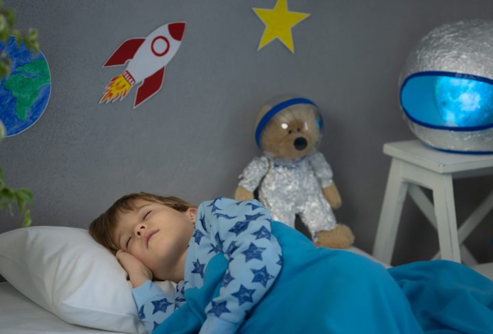 Happy child sleeping with toy rocket and teddy bear. Kid pretend to be astronaut. Imagination and children dream concept